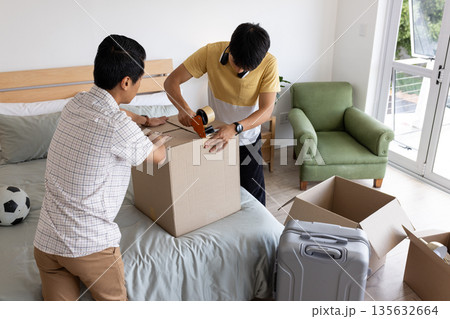 Two men packing cardboard box on bed, preparing for move with suitcase nearby 135632664