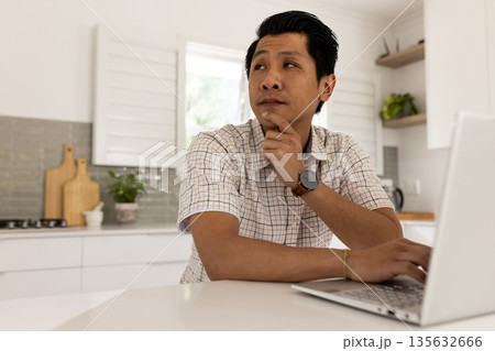 Asian man in kitchen using laptop, thinking deeply and planning work Asian man in kitchen using laptop, thinking deeply and planning work 135632666