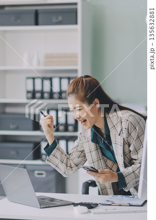 Determined asian businesswoman works late in a modern office, confidently analyzing data on her laptop, embodying success and professionalism with a smile 135632881