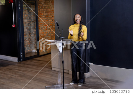 African American woman in mustard top clapping behind clear lectern with microphone in event space 135633756