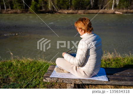 Woman playing a steel tongue drum in nature with a mountain river and Altai landscape in the background. Concept of harmony, mindfulness, and connection with nature Woman playing a steel tongue drum in nature with a mountain river and Altai landscape in the background. Concept of harmony, mindfulness, and connection with nature 135634318