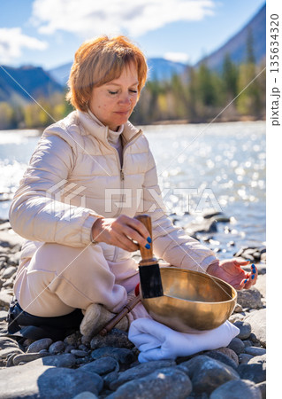 Woman performing a sound ritual with a Tibetan singing bowl by the Altai river. Concept of feminine energy, sacred vibration, and shamanic connection to the Earth Woman performing a sound ritual with a Tibetan singing bowl by the Altai river. Concept of feminine energy, sacred vibration, and shamanic connection to the Earth 135634320