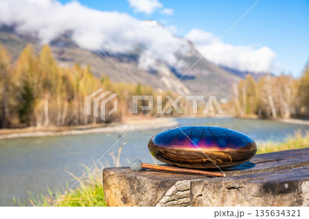 Steel tongue drum and mallets resting on a wooden bench with mountains, river, and forest in the background. Concept of sacred stillness, sound meditation, and harmony with nature 135634321
