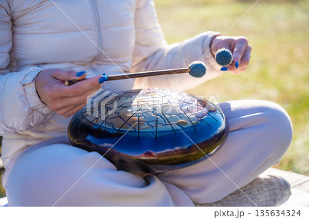 Close-up of hands playing a steel tongue drum with mallets against the backdrop of Altai mountains and river. Concept of sacred sound, touch of nature, and mindful musical ritual 135634324