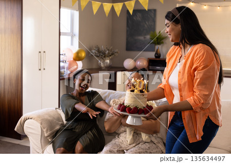 Woman presenting birthday cake with candles to joyful friends at home celebration Woman presenting birthday cake with candles to joyful friends at home celebration 135634497