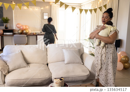 Mother holding baby in living room, celebrating with family during daytime Mother holding baby in living room, celebrating with family during daytime 135634517