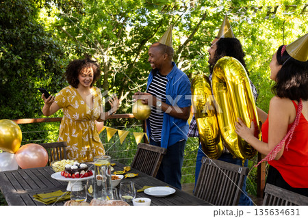 Friends celebrating birthday outdoors with cake and balloons, wearing party hats Friends celebrating birthday outdoors with cake and balloons, wearing party hats 135634631