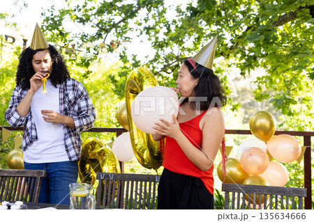 Friends celebrating outdoors, wearing party hats, blowing balloons and party blowers 135634666