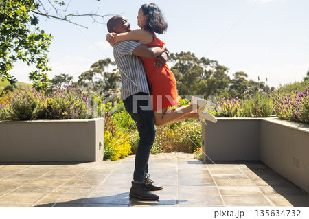 Couple joyfully embracing outdoors, surrounded by vibrant garden on sunny day Couple joyfully embracing outdoors, surrounded by vibrant garden on sunny day 135634732
