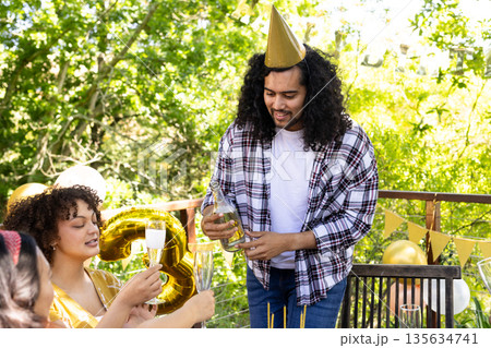 Man pouring champagne at outdoor birthday party with friends, wearing party hat Man pouring champagne at outdoor birthday party with friends, wearing party hat 135634741