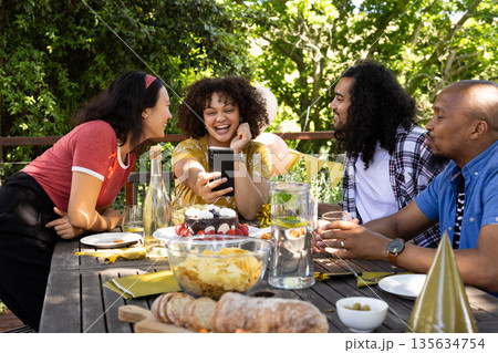 Celebrating outdoors, friends laughing and taking selfie at festive table with snacks Celebrating outdoors, friends laughing and taking selfie at festive table with snacks 135634754