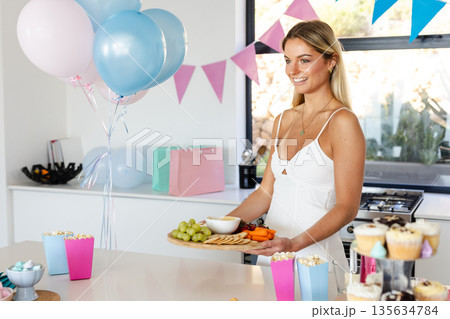Smiling woman preparing snacks for party with balloons and decorations at home Smiling woman preparing snacks for party with balloons and decorations at home 135634784