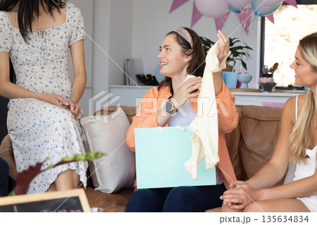 Women celebrating baby shower, smiling and opening gifts on cozy living room couch Women celebrating baby shower, smiling and opening gifts on cozy living room couch 135634834