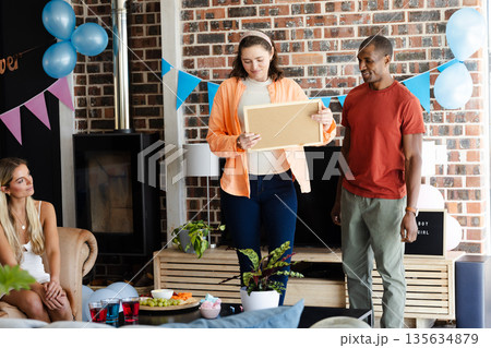 Friends celebrating at home, woman holding gift while others smiling and chatting 135634879