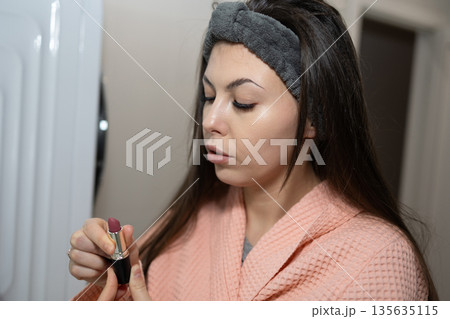 Woman Applies Lipstick in Bathroom While Wearing a Bathrobe in the Evening 135635115