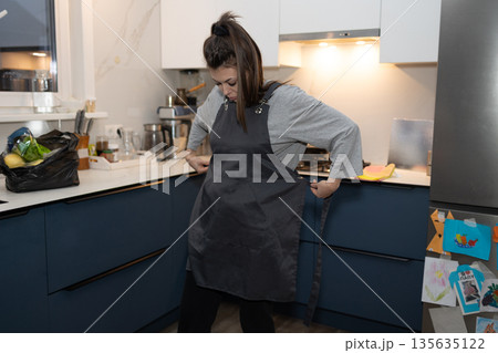 Woman Adjusting Apron in Bright Modern Kitchen Before Preparing Food Woman Adjusting Apron in Bright Modern Kitchen Before Preparing Food 135635122