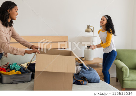 Mother and daughter packing clothes into cardboard box in bedroom, smiling together Mother and daughter packing clothes into cardboard box in bedroom, smiling together 135635534
