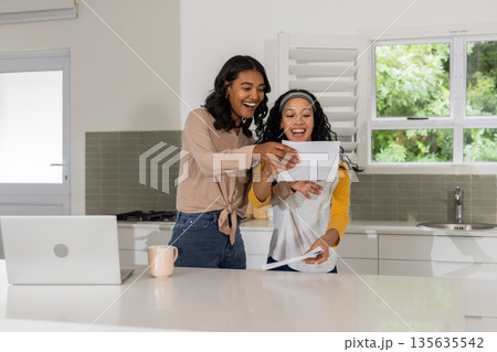 Mother and daughter joyfully reading letter in modern kitchen with laptop nearby Mother and daughter joyfully reading letter in modern kitchen with laptop nearby 135635542