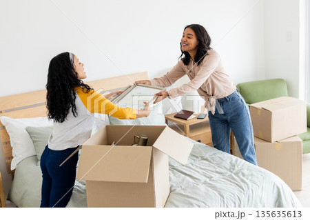 Two women unpacking boxes on bed, smiling and organizing new home together Two women unpacking boxes on bed, smiling and organizing new home together 135635613