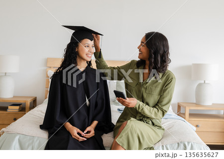 Proud mother adjusting daughter's graduation cap while sitting on bed at home 135635627