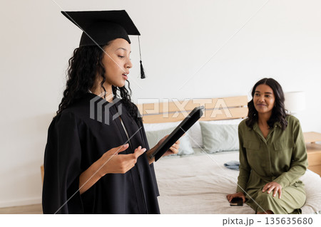Asian graduate in cap and gown reading speech at home with proud mother watching 135635680