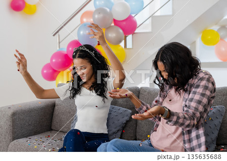 Celebrating at home, two women joyfully throwing confetti with colorful balloons Celebrating at home, two women joyfully throwing confetti with colorful balloons 135635688
