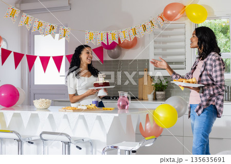 Celebrating birthday at home, two women enjoying cake and snacks with balloons 135635691