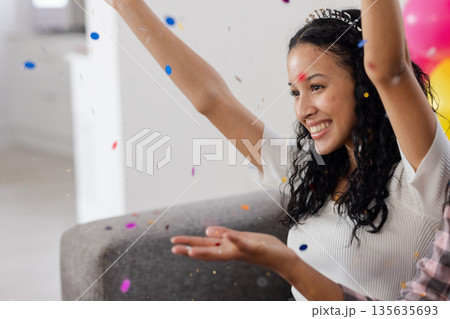 Joyful woman celebrating with confetti, smiling and raising arms at home Joyful woman celebrating with confetti, smiling and raising arms at home 135635693