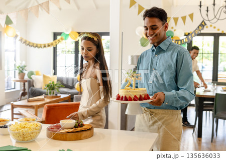 Young man carrying birthday cake while woman arranges snacks at home party 135636033