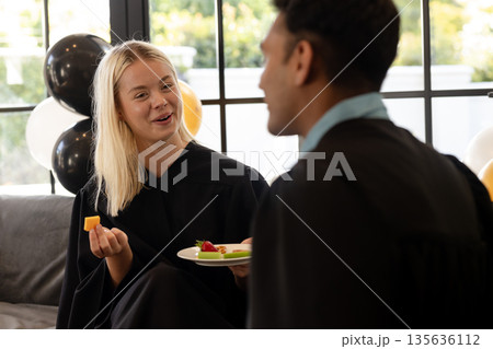 Graduates in gowns enjoying snacks and conversation at home celebration 135636112