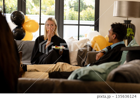 Graduates in gowns relaxing on sofa, enjoying snacks at home celebration Graduates in gowns relaxing on sofa, enjoying snacks at home celebration 135636124