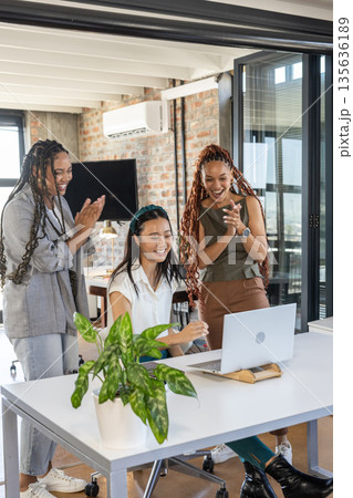 Team celebrating success at office, three women clapping and smiling at laptop 135636189