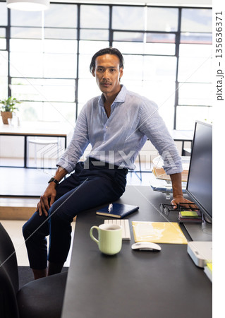 Confident businessman sitting on desk in modern office, looking at camera 135636371