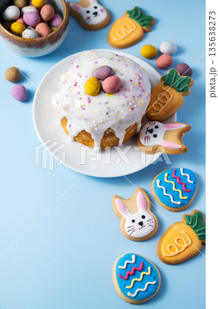 Easter cake with white icing glaze, chocolate mini eggs and easter cookies: bunny, carrots and blue egg cookies. Top view, blue background. Easter cake with white icing glaze, chocolate mini eggs and easter cookies: bunny, carrots and blue egg cookies. Top view, blue background. 135638273