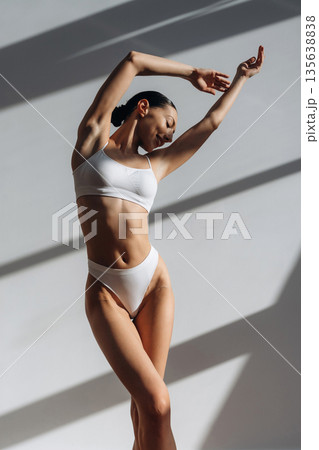 Against the white wall, standing and posing. Beautiful young woman with slim body type is indoors Against the white wall, standing and posing. Beautiful young woman with slim body type is indoors 135638838