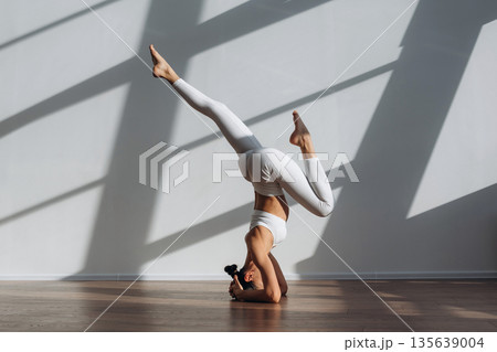 Headstand, doing exercise. Beautiful young woman with slim body type is indoors Headstand, doing exercise. Beautiful young woman with slim body type is indoors 135639004