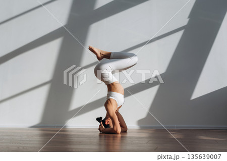 Headstand, doing exercise. Beautiful young woman with slim body type is indoors Headstand, doing exercise. Beautiful young woman with slim body type is indoors 135639007