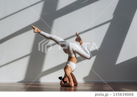 Headstand, doing exercise. Beautiful young woman with slim body type is indoors Headstand, doing exercise. Beautiful young woman with slim body type is indoors 135639008