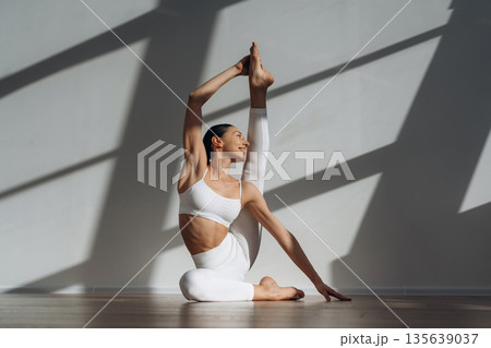 Model is doing yoga exercises. Beautiful young woman with slim body type is indoors Model is doing yoga exercises. Beautiful young woman with slim body type is indoors 135639037