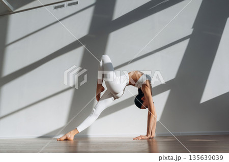 Model is doing yoga exercises. Beautiful young woman with slim body type is indoors Model is doing yoga exercises. Beautiful young woman with slim body type is indoors 135639039