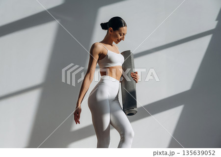 Gray yoga mat in hand. Beautiful young woman with slim body type is indoors 135639052