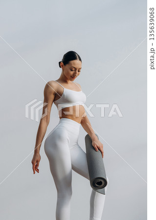 Fitness trainer is holding yoga mat. Beautiful young woman with slim body type is indoors 135639068