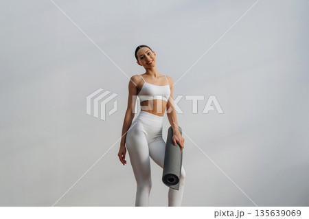Fitness trainer is holding yoga mat. Beautiful young woman with slim body type is indoors 135639069