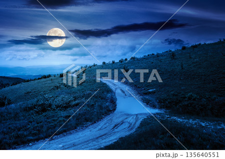 country dirt road through mountains on a summer night. scenic view of rolling hills under dark sky with clouds in full moon light. beautiful alpine meadow for finance investment or abstract concepts 135640551