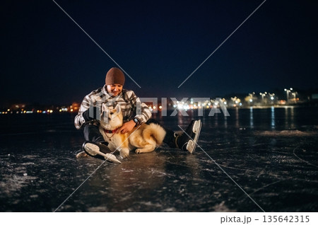 Resting by sitting on frozen lake. Man with Akita Inu dog is on the ice with skates in winter Resting by sitting on frozen lake. Man with Akita Inu dog is on the ice with skates in winter 135642315