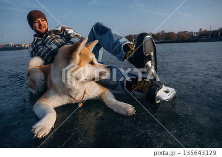 Just lying down and resting. Man with Akita Inu dog is on the ice with skates in winter Just lying down and resting. Man with Akita Inu dog is on the ice with skates in winter 135643129