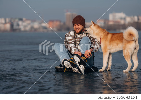 Ties shoelaces, sitting. Man with Akita Inu dog is on the ice with skates in winter 135643151
