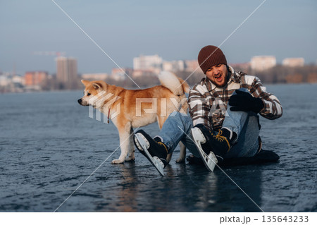 Pain in the leg, fell. Man with Akita Inu dog is on the ice with skates in winter Pain in the leg, fell. Man with Akita Inu dog is on the ice with skates in winter 135643233