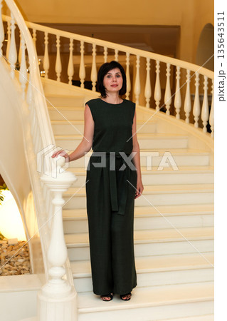 A mature, slender woman in an elegant evening dress standing on a beautiful marble staircase 135643511