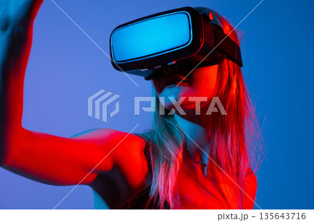 Close-up of woman wearing VR headset and raising arm in virtual space, immersive technology with neon red and blue lighting Close-up of woman wearing VR headset and raising arm in virtual space, immersive technology with neon red and blue lighting 135643716